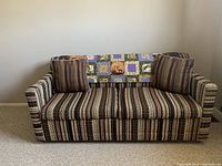 Photo of a retro striped fabric hide-a-bed couch with two striped cushions and a patchwork quilt on the backrest, showcasing overall design and condition.