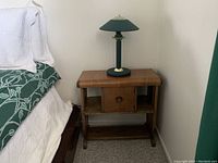 Photo of wooden side table with central drawer, lower shelf, and green desk lamp on top next to a bed with green and white bedding.