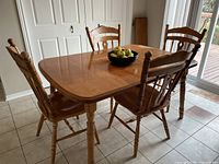 Photo showing the rectangular wooden dining table with four matching chairs around it. The table has a medium-tone wood finish with rounded edges and turned legs.