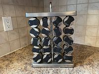Front view of the stainless steel spice rack holding 16 round spice containers with black lids on a kitchen countertop.