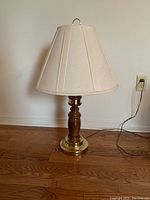 Table lamp composed of a wooden turned body with brass base, beige fabric shade, standing on wood floor near wall outlet