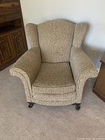 Winged armchair upholstered in woven textured fabric with caster wheels on its wooden legs situated indoors near wooden furniture.