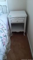 Front and side view of the white wooden bedside table next to a bed with floral bedding, showing drawer and open shelf.