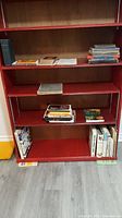 Red bookshelf with multiple shelves holding assorted books including novels, history, and gardening books.