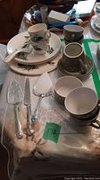 Photo showing two Jon Anton ironstone cups and saucers, cream and sugar pieces placed near a ceramic server and plates with floral design. Silver-tone lifters and cutting utensil with decorative handles positioned in front.