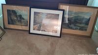 Three wood framed landscape pictures leaning against wall on carpeted floor. Variety of wood frame colors and sizes noted.