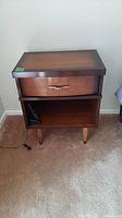 Front view of a Mid-Century Modern bedside table showing drawer with gold-tone handle and open shelf below, placed on carpet.