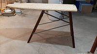 Side angled view of the wood framed ironing board with fabric cover showing the wooden legs and metal support bars, situated on a concrete floor in a basement.