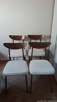 Front view of four Mid-Century Modern dining chairs with brown wooden backs and white upholstered seats on metal legs.
