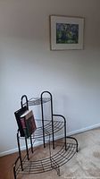 Metal semi circle-shaped plant/book stand with black wire three shelves and vertical handles, displaying two books on the middle shelf, shown from front angle.