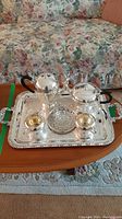 Photo showing full silverplate tea service set on wooden coffee table with floral sofa backdrop, including tray, pots, and accessories