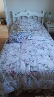 White wooden twin bed made with floral comforter and some linens folded on top