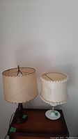 Full view of two lamps on wooden surface showing one milk glass hob nail lamp with white pom-pom trimmed shade and one mid-century wood lamp with beige scalloped shade.