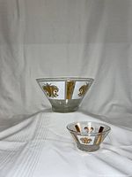 Two conical glass bowls, one large and one small, each decorated with gold and white vertical panels featuring fleur-de-lis motifs and rectangles. Both bowls are set on a white fabric background.