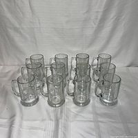 Photo of all 12 identical clear glass beer mugs arranged in rows on fabric background.