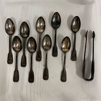 Top view of eight metal spoons and one pair of metal tongs placed on white textured fabric, showing tarnishing and wear on spoons and tongs.