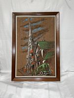 Framed handpainted canvas artwork showing a street scene with a tall church spire, buildings with green roofs, a green vehicle, several figures, and a cloudy sky with blue accents. Medium brown wooden frame with white inner trim.