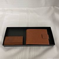 Photo of the two leather items side by side in a black tray on a white textured fabric surface. Shows the full external appearance of the wallet and key holder.