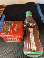 Red Coca Cola tin and Coca Cola bottle-shaped thermometer together on black surface
