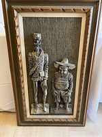 Framed ceramic wall art with Don Quixote and Sancho Panza ceramic figures mounted on textured fabric background.