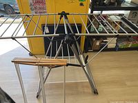 Metal tripod-style clothes drying rack with multiple rods for drying clothes, partially unfolded and standing on floor.