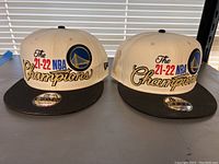 Two Golden State Warriors 21-22 NBA Champions white and black snapback hats placed side by side on a surface