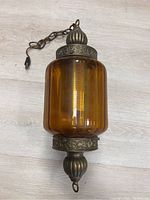 Full view of the hanging lamp showing amber glass shade and metal top and bottom finials with chain and electrical cord.