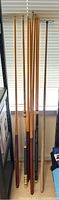 Photo showing seven wooden pool cues standing upright in a row, illustrating variety in length and diameter.