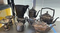 Wide view of silverplated collection showing various tea and coffee service pieces including pitchers, teapots, and bowls on shelf with light from window behind.