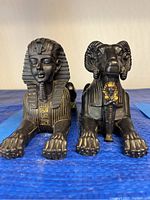 Front view of the black Sphinx statue and the Ram statue placed side by side on a blue surface.