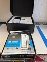 Boxed Arris SURFboard SB6141 modem standing on a surface with black open box behind showing camera and accessories inside.
