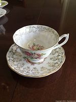 Bone china teacup displaying elegant floral pink rose pattern with gold trim on rim and handle, sitting on matching saucer with intricate floral and gold design.