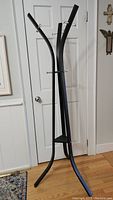 Front view of a sturdy black aluminum coat rack with curved vertical poles and three hooks, standing on wooden floor against a white door and wall.
