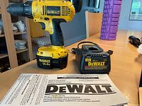 Yellow DeWalt cordless drill with battery attached, battery charger, and instruction manual lying on a wooden table.
