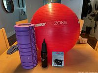 Large red inflated exercise ball with 'GOZONE' branding, purple foam roller, black pump, and new black whistle on lanyard in package, arranged on wood floor with chairs in background.