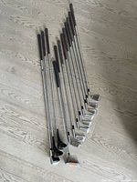 Full view of 12 left hand golf clubs laid out on floor showing club heads, shafts and grips.