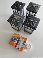 Four black plastic mosquito protection canisters with lattice tops and adjustable tabs, accompanied by two OFF! brand repellent refill packs in orange packaging and one white tealight candle, all placed on a white surface.