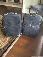 Two identical black nylon suitcases with multiple front compartments standing upright on wood floor near carpet and chair.