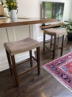 Two wooden bar stools with beige upholstered seats featuring nailhead trim, placed next to a kitchen counter. The upholstery is worn and needs reupholstering.