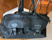 Front view of the black leather Roots weekender bag showing two exterior buckle compartments and embossed logo