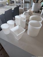 Photo showing various white BPA-free plastic cheese molds in different shapes including cylindrical and round molds with multiple perforations for drainage. Also visible is a round drip tray and a folded cheese cloth.