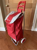 Front-side view of the red lightweight aluminum framed shopping trolley showing the flap with branding and the drawstring bag.