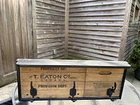 Rustic wall mounted coat rack made from wooden box panels with black frame and three metal hooks, showing box text and marks.