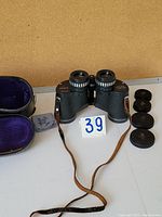 Full front view of black textured binoculars with attached aged brown leather strap, lens caps, original case open showing purple interior, and cleaning cloth beside them