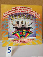 Front cover of The Beatles Magical Mystery Tour vinyl LP, colorful psychedelic artwork with band in costumes and track listing.