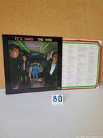 Another angle front photo of The Who "It's Hard" album and white inner sleeve with printed lyrics or notes, showing vinyl and sleeve condition.