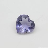 Close-up photo of a heart-shaped violet-blue Iolite gemstone placed on a white background highlighting its facets and color.