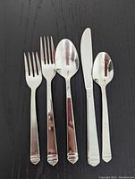12 place settings of Lagostina Indonesia stainless steel flatware including serrated knives, soup spoons, dinner forks, salad forks, and dessert spoons arranged in a row on black surface.