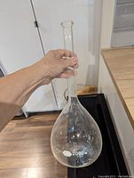 Photo of a clear glass laboratory beaker with a long narrow neck and round base, hand held, showing 2000 ml capacity mark and Kimax brand.