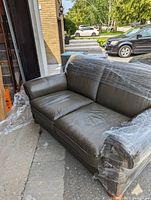 Front-left angled view of loveseat wrapped in plastic showing overall shape, arm style, cushions